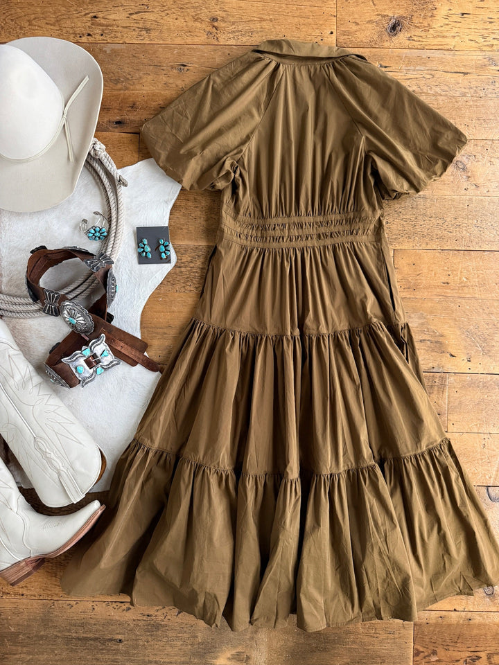 The Monterey Bronze Olive Puff Sleeve Dress by Savannah Sevens western life{&}style is laid flat on a wooden floor, surrounded by a white cowboy hat, white boots, a coiled rope belt, a leather belt with a turquoise buckle, and turquoise jewelry.