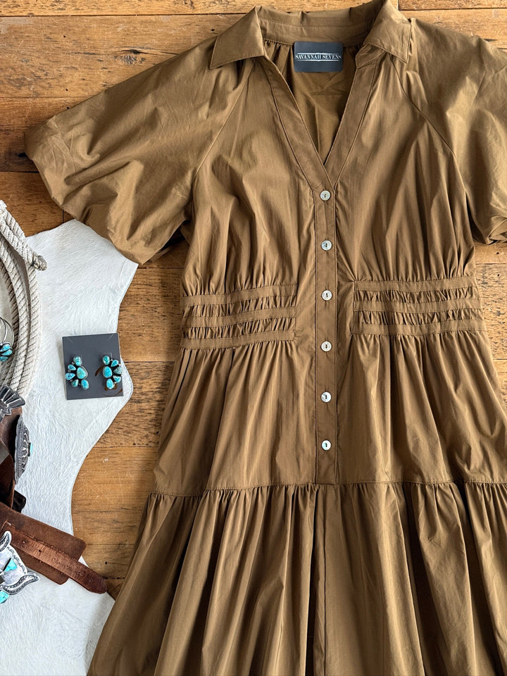 The Monterey Bronze Olive Puff Sleeve Dress by Savannah Sevens western life{&}style, a short-sleeve button-up cotton blend dress with a tiered skirt, is laid on a wooden surface. Nearby are turquoise earrings, a cowhide purse, and part of a brown leather belt.