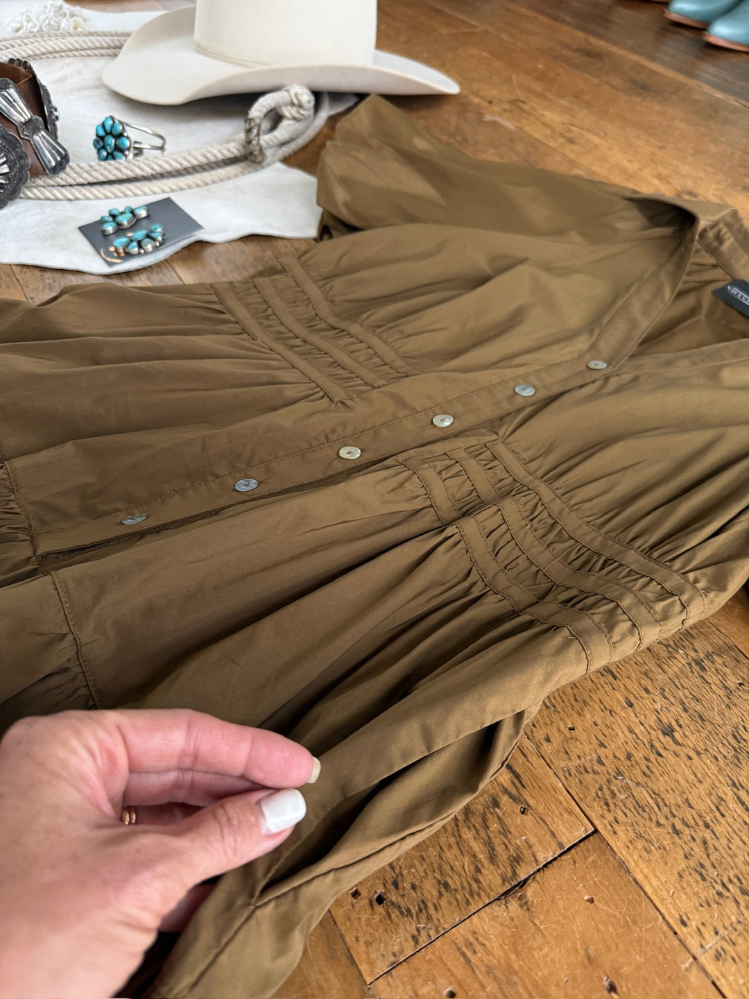 A hand holds the sleeve of the Monterey Bronze Olive Puff Sleeve Dress by Savannah Sevens western life{&}style, laid out on a wooden floor. In the background are a white hat, turquoise jewelry, a belt, and brown western boots.