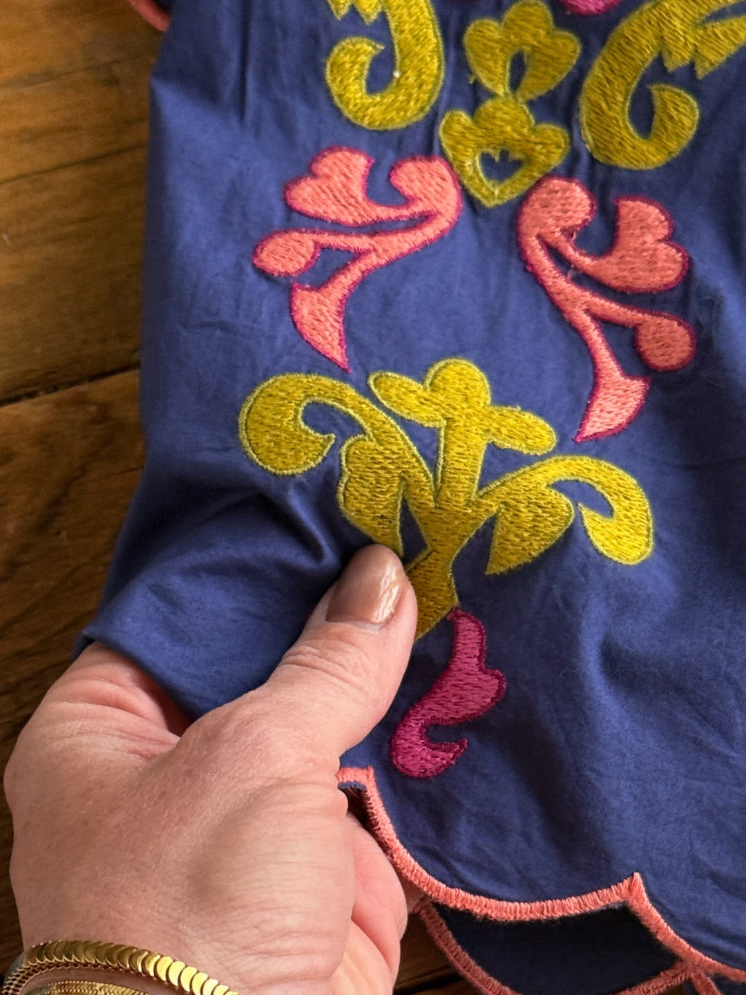A hand with  a gold bracelet holds The Montevello, a blue floral embroidered top by Savannah Sevens western life{&}style, featuring yellow and pink details, displayed on a wooden surface.