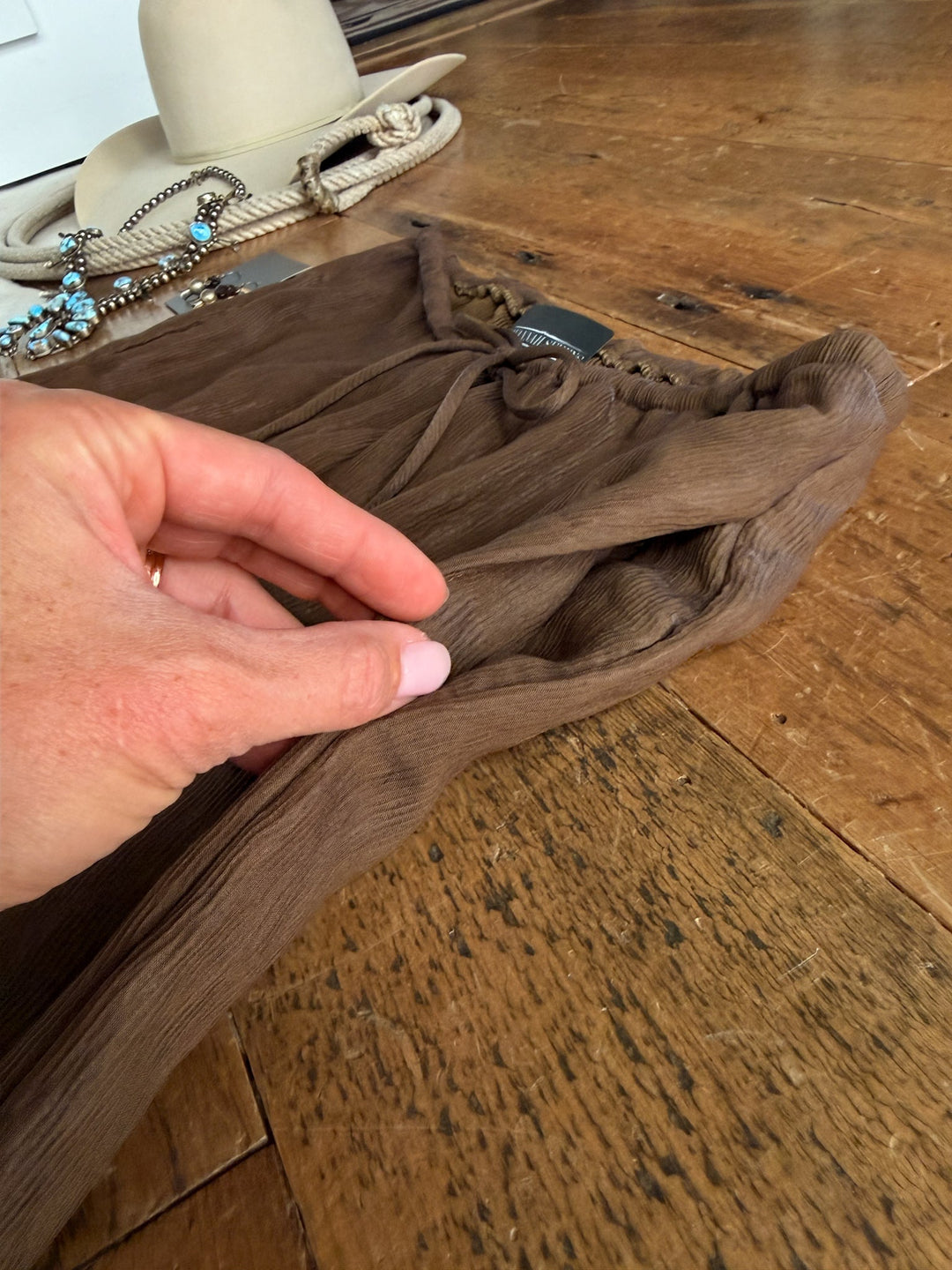 A hand lifts the fabric of the Montezuma Olive Brown Wide Leg Pants Set by Savannah Sevens western life{&}style on a wooden floor. In the background, a beige hat, beaded turquoise jewelry, and a coiled rope are visible.