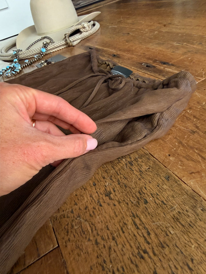 A hand lifts the fabric of the Montezuma Olive Brown Wide Leg Pants Set by Savannah Sevens western life{&}style on a wooden floor. In the background, a beige hat, beaded turquoise jewelry, and a coiled rope are visible.