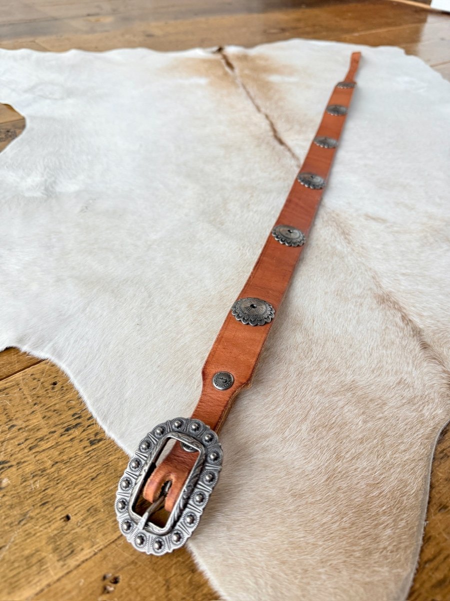 The Monticello Belt by Savannah Sevens western life{&}style features cognac leather with southwest silver conchos and a large engraved interchangeable buckle, shown on animal hide over a wooden floor.