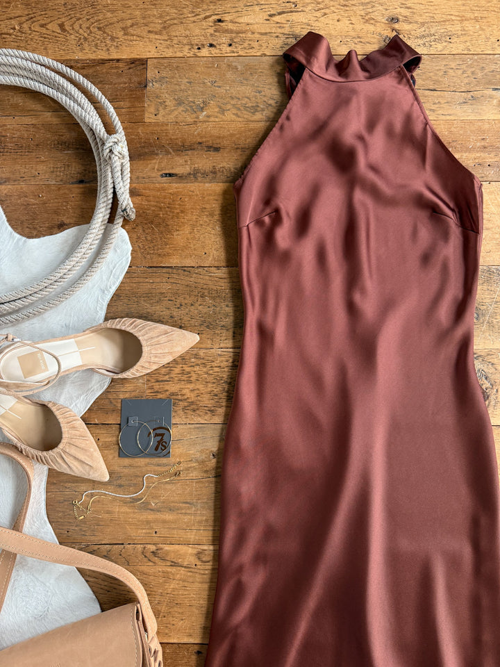 The Montrose Chestnut Halter Neck Dress by Savannah Sevens western life{&}style is displayed on a wooden floor alongside beige heels, a white textured bag with thick handles, a tan purse, and gold jewelry.