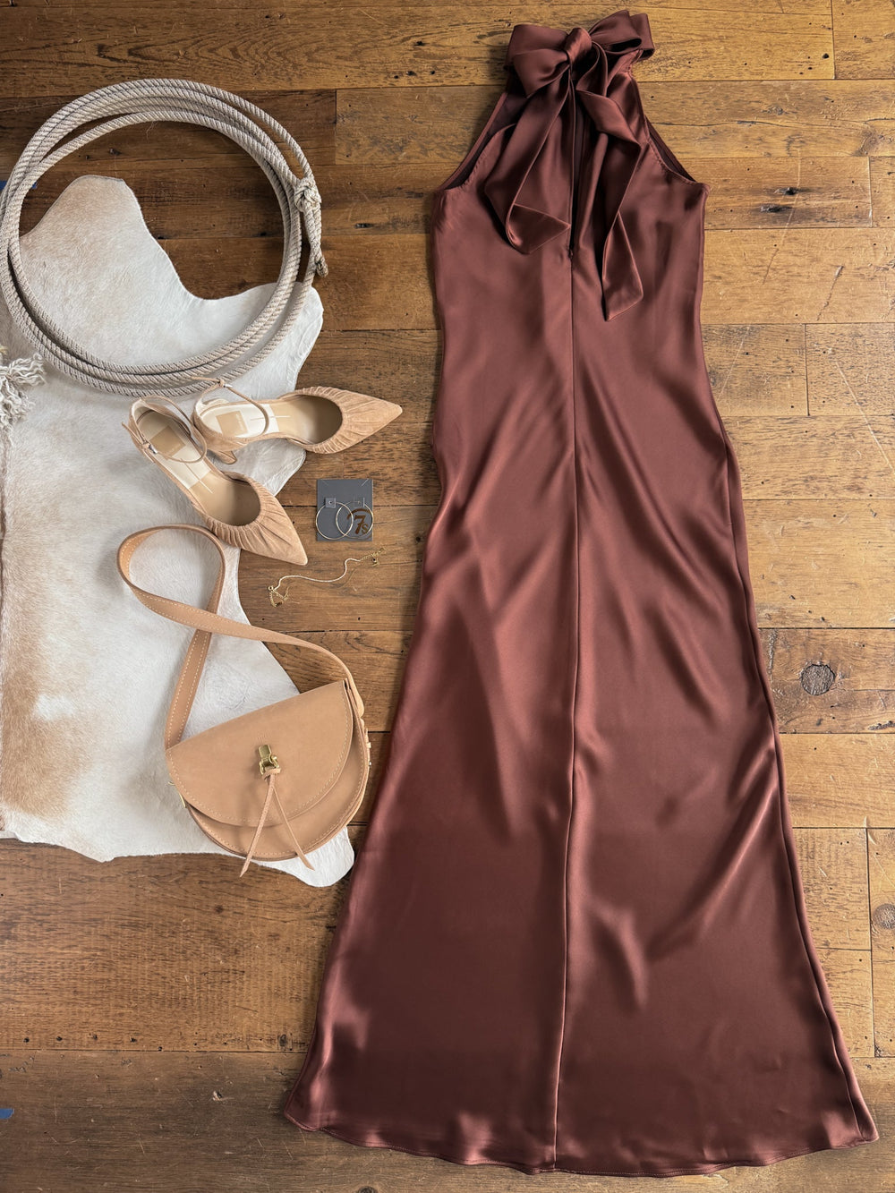 The Montrose Chestnut Halter Neck Dress by Savannah Sevens western life{&}style is styled with tan heels, a tan purse, silver earrings, and displayed on a cowhide rug atop a wooden floor.