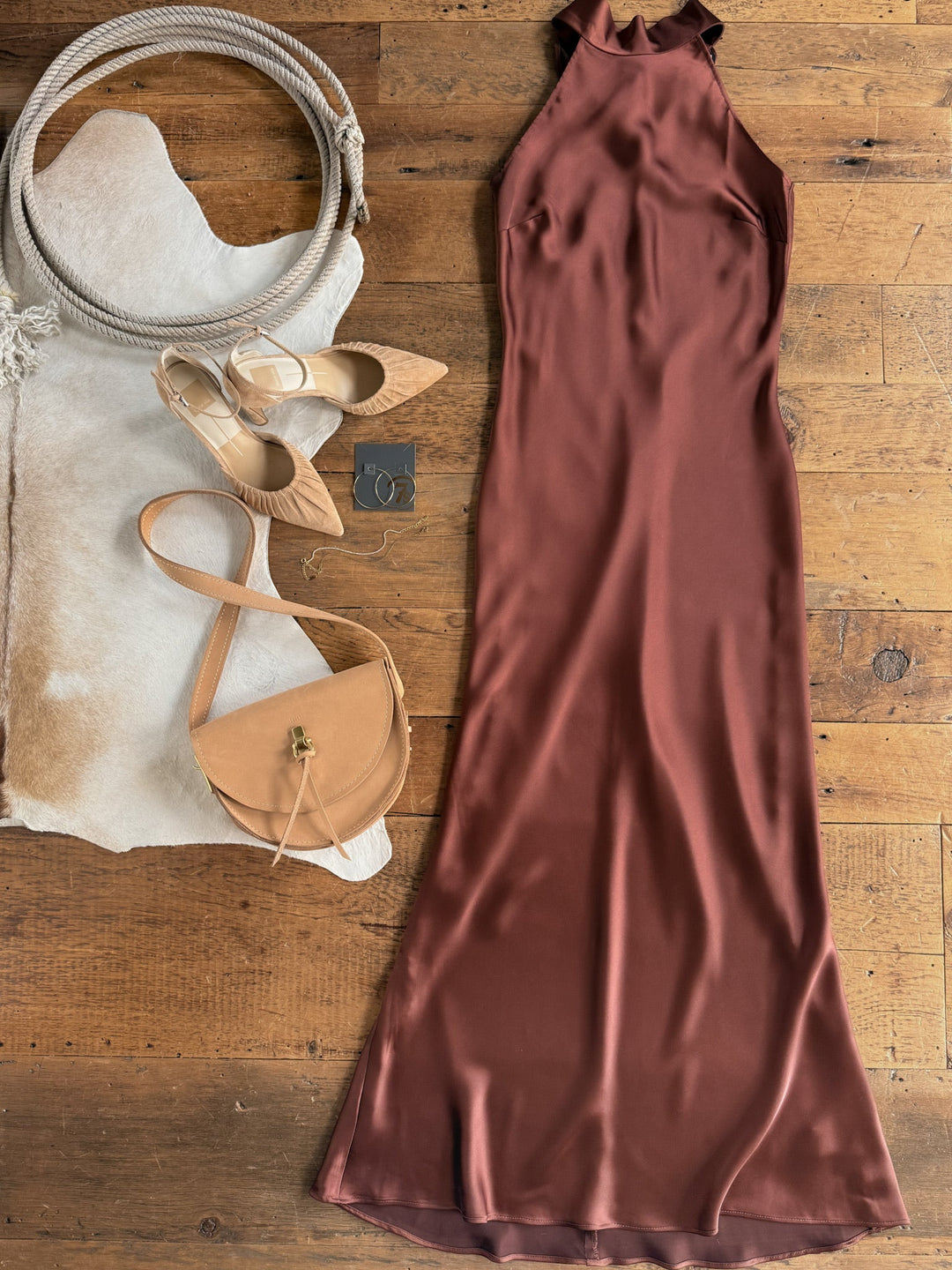 The Montrose Chestnut Halter Neck Dress by Savannah Sevens western life{&}style, paired with tan pointed heels, a tan crossbody purse, and gold hoop earrings, is arranged on a wooden floor and white cowhide.