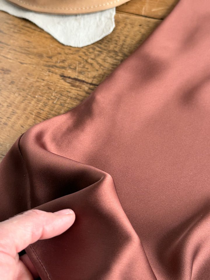 A hand holds the edge of the Savannah Sevens western life{&}style Montrose Chestnut Halter Neck Dress, which is laid out on a wooden surface with part of a tan object visible in the background.