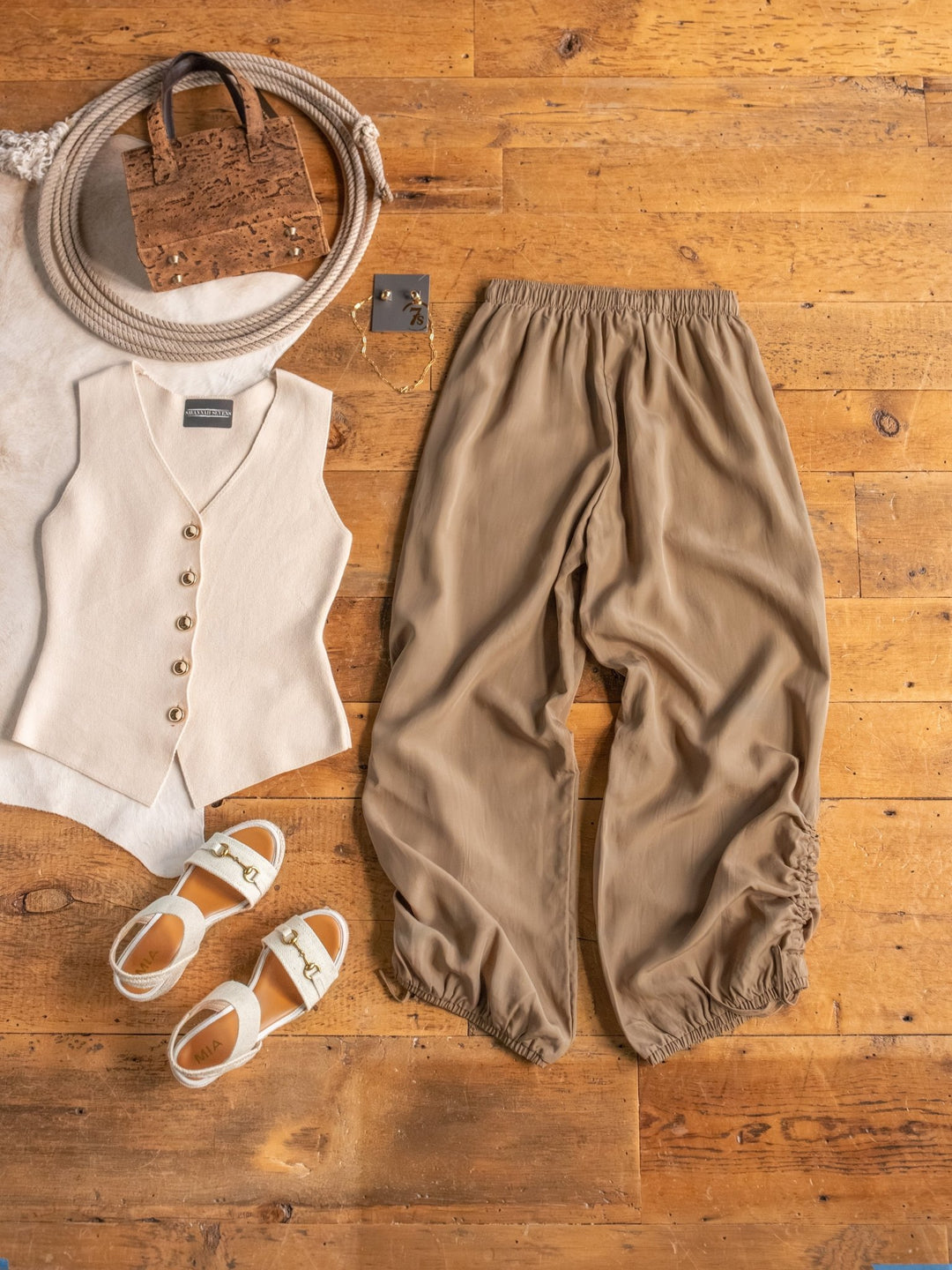 The Moroa by Savannah Sevens western life{&}style—a beige vest with gold buttons, linen blend wide leg pants, white sandals, bag, and silver square earrings—arranged on a wooden floor.
