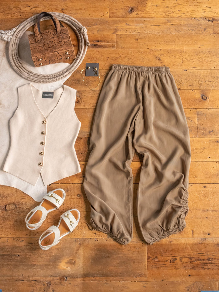 The Moroa by Savannah Sevens western life{&}style—a beige vest with gold buttons, linen blend wide leg pants, white sandals, bag, and silver square earrings—arranged on a wooden floor.