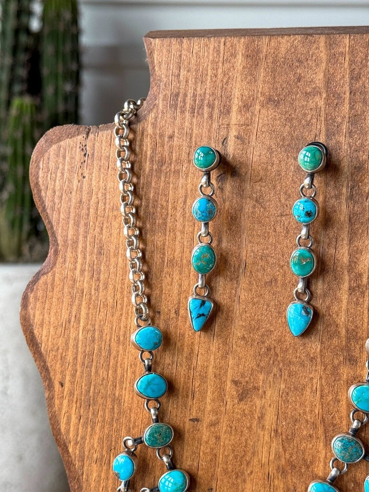 A wooden jewelry display showcases the Morado Necklace & Earrings Set by Savannah Sevens western life&style, featuring Sonoran turquoise and sterling silver with round and teardrop stones. A blurred cactus appears in the background.