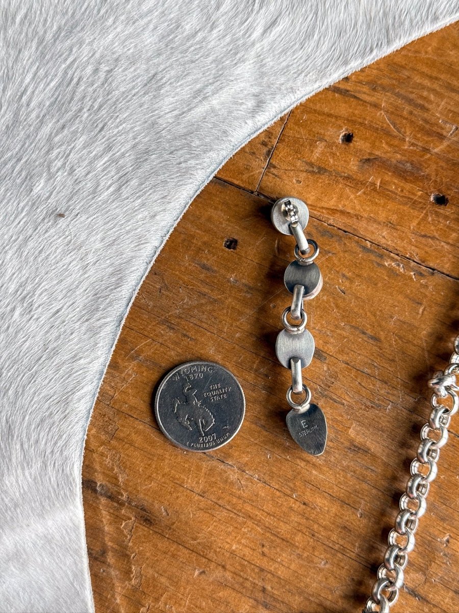 The Morado Necklace & Earrings Set by Savannah Sevens western life&style is shown: silver earrings with three linked segments rest beside a US quarter on wood, along with a sterling chain and light cowhide.
