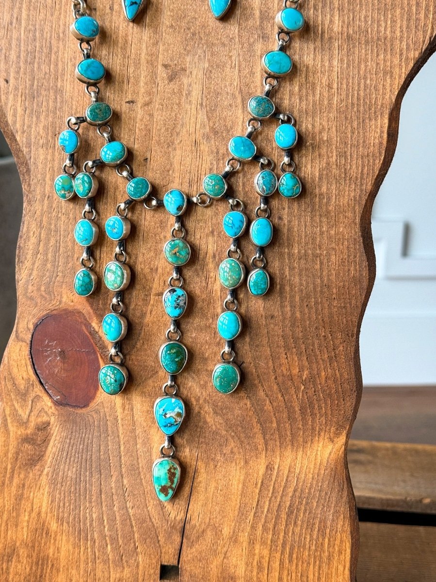 The Morado Necklace & Earrings Set by Savannah Sevens western life&style features sterling silver with oval and teardrop Sonoran turquoise stones arranged in dangling rows, displayed on a wooden necklace stand.