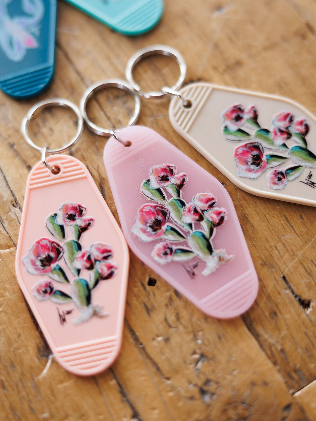 Three Savannah Sevens western life{&}style Motel Keychains with painted cactus flower designs in pastel pink and beige rest on a wooden surface. These vintage-inspired keychains are perfect as stylish purse accessories.