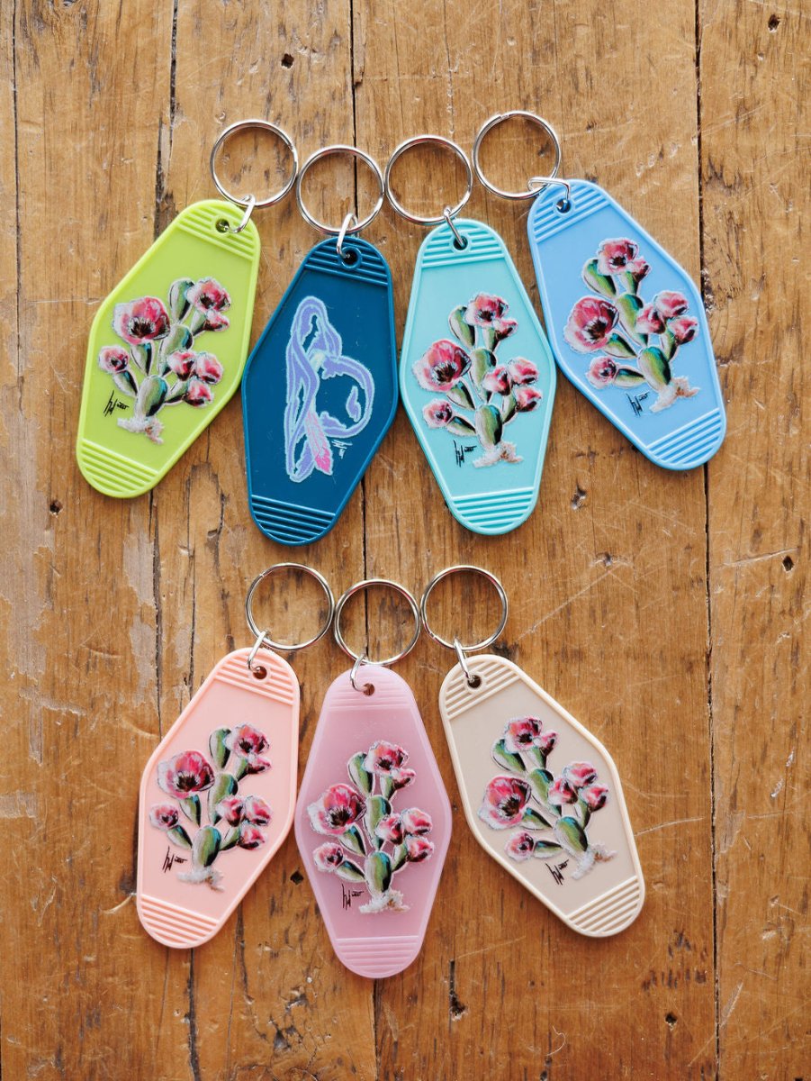 Eight Savannah Sevens western life{&}style Motel Keychains—seven in pastel cactus floral designs and one blue with a bird graphic—are displayed on rustic wood, perfect as purse accessories.