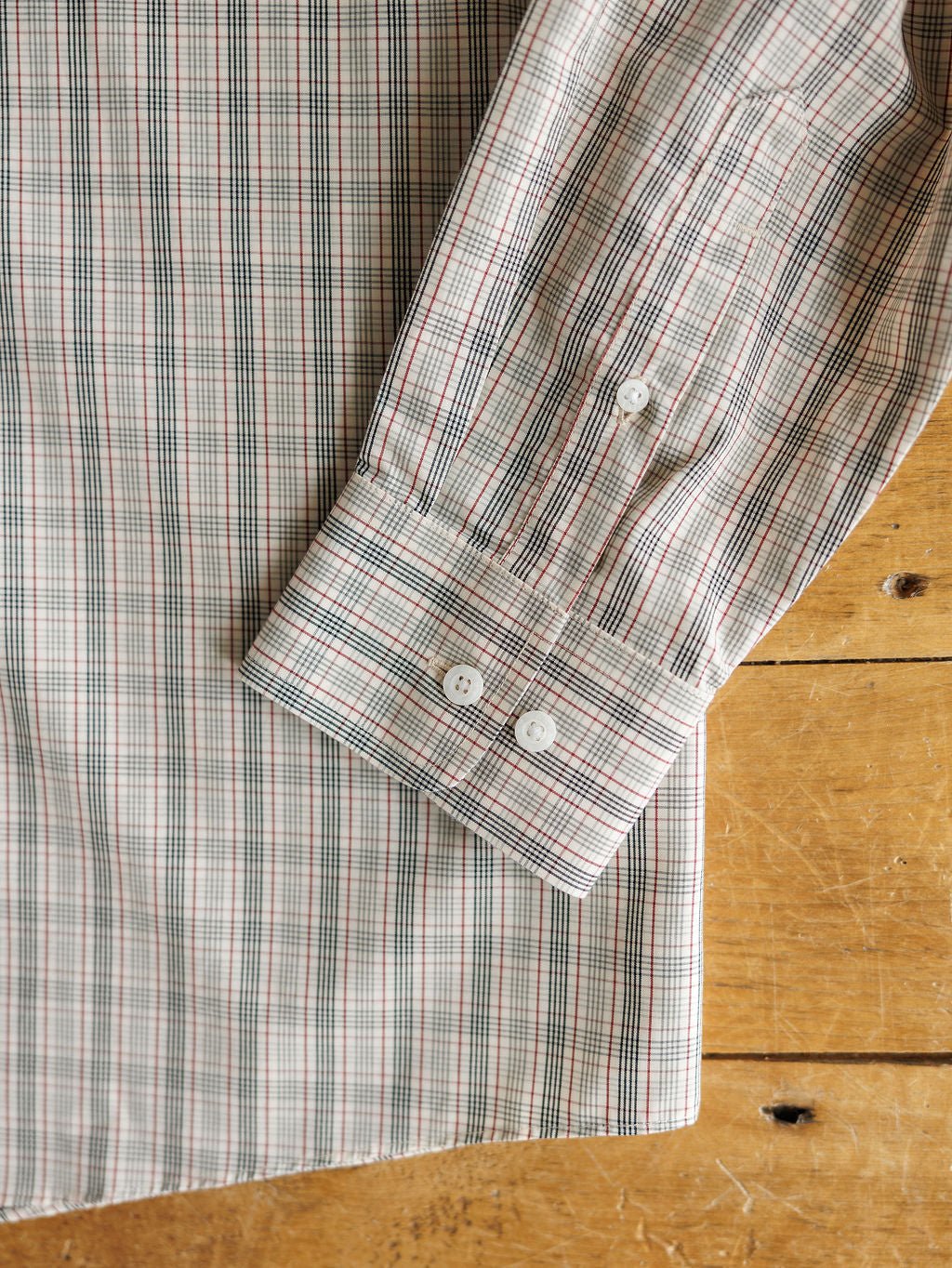 A close-up of the Murtaugh Ariat Men's Natural Plaid Button Up by Savannah Sevens western life{&}style, showing its red, green, and white striped plaid long sleeves with a cuff featuring two white buttons, laid flat on wood.