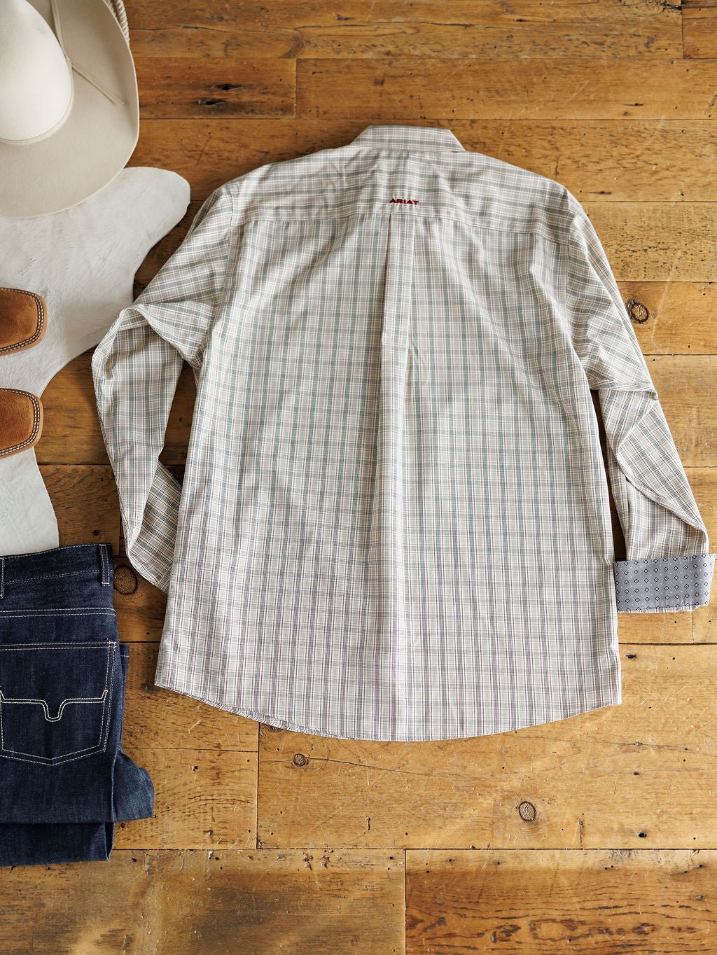 The Murtaugh Ariat Men's Natural Plaid Button Up by Savannah Sevens western life{&}style is laid flat on a wooden floor, styled with a white cowboy hat, tan boots, and folded dark jeans. The back of the shirt is shown.