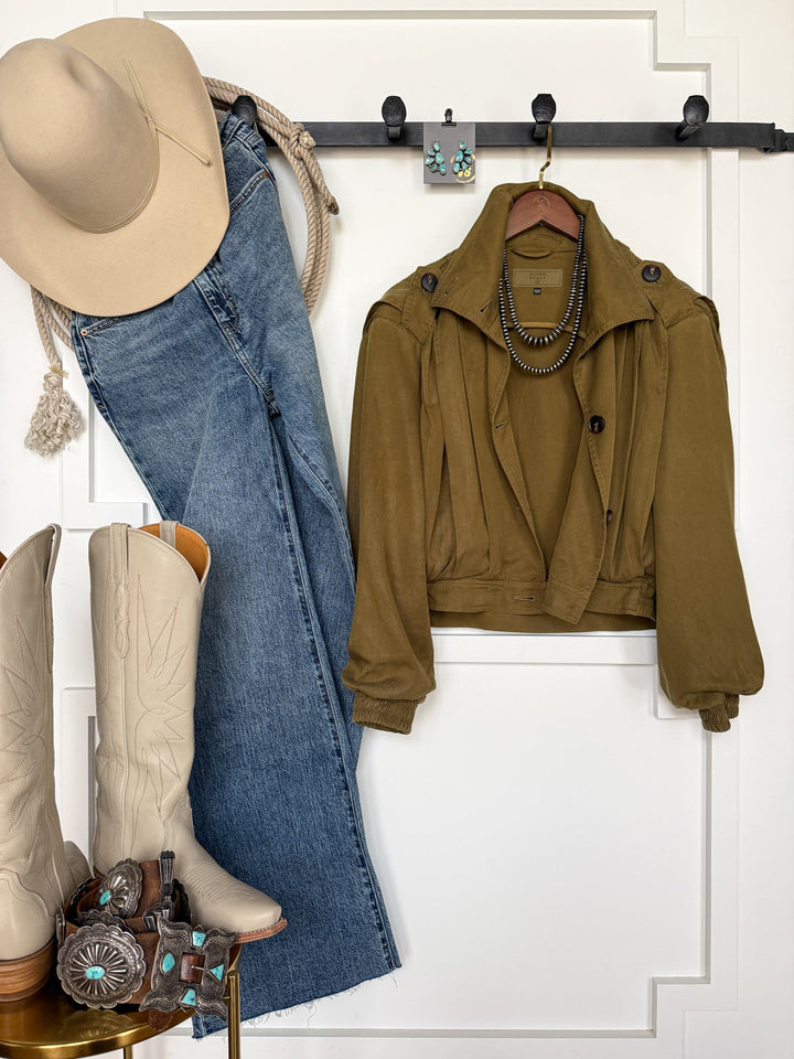 A Western outfit arranged on a wall features a beige cowboy hat, blue jeans, the Natoma Olive Button Up Jacket by Savannah Sevens western life{&}style on a hanger, tan cowboy boots, turquoise jewelry, and a coiled rope—all displayed on a black wall rack against a white background.