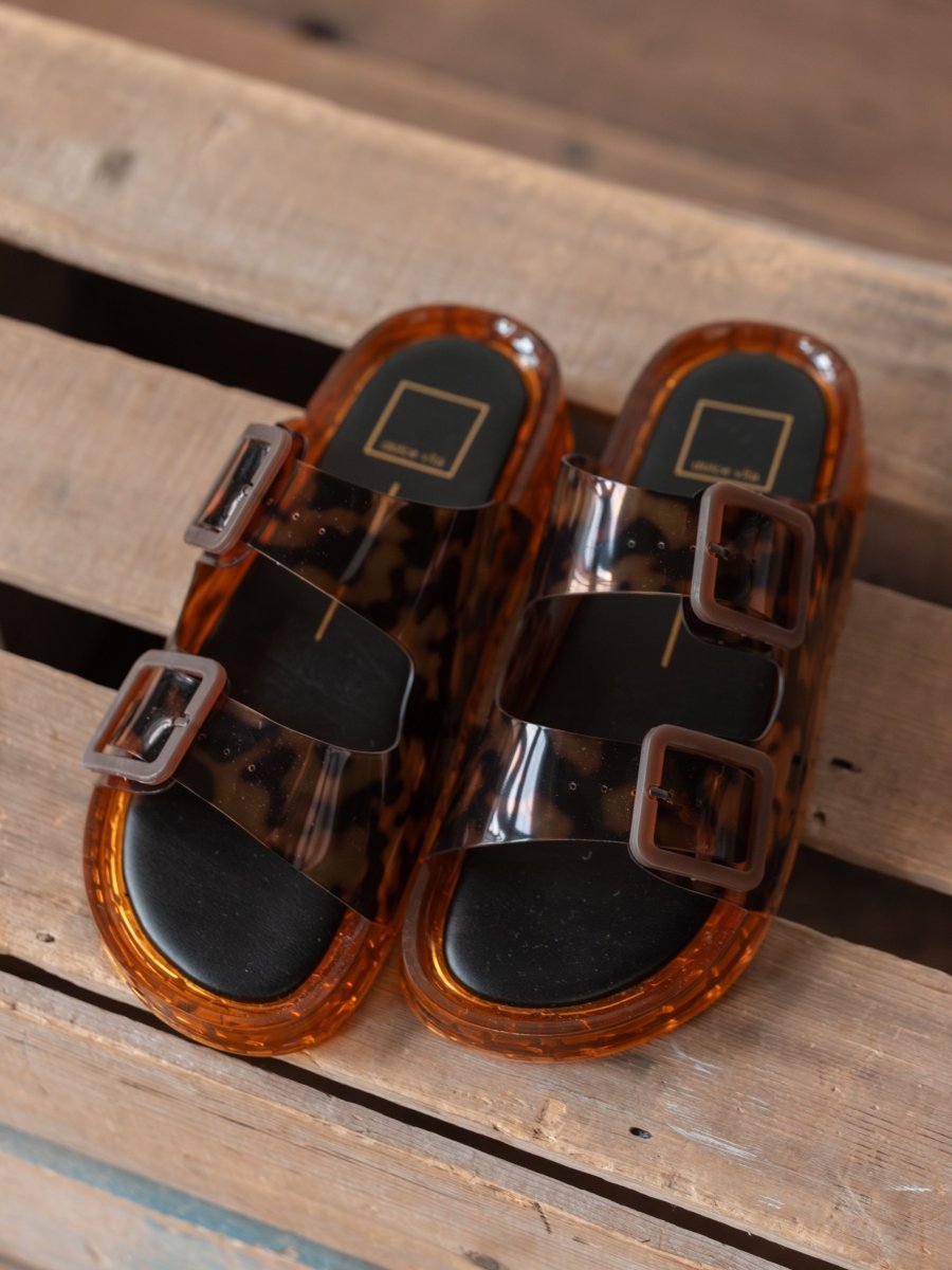 The Navarro by Savannah Sevens western life{&}style: stylish tortoise slip-on sandals with transparent brown straps, large rectangular buckles, adjustable cushioned footbed, and orange translucent soles, displayed on a wooden surface.