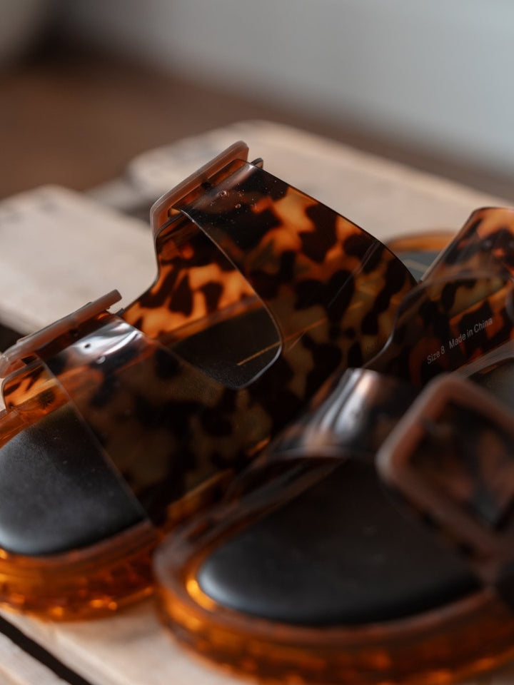 Close-up of The Navarro sandals by Savannah Sevens western life{&}style, featuring adjustable transparent brown straps, dark cushioned footbeds, and chunky soles on a light wooden surface.
