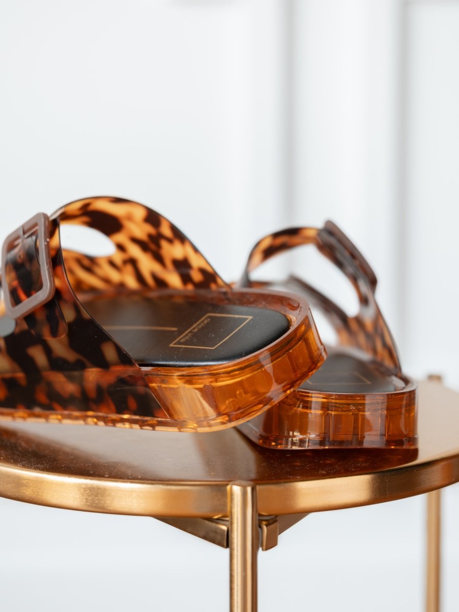 Close-up of The Navarro slip-on sandals by Savannah Sevens western life{&}style, featuring adjustable straps and a cushioned footbed, displayed on a round metallic gold table with a blurred white background.