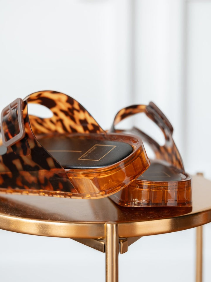 Close-up of The Navarro slip-on sandals by Savannah Sevens western life{&}style, featuring adjustable straps and a cushioned footbed, displayed on a round metallic gold table with a blurred white background.