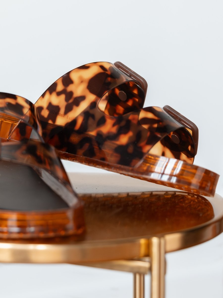 A close-up of The Navarro slip-on sandals by Savannah Sevens western life{&}style, featuring brown and black tortoise adjustable straps, displayed on a round gold metallic table against a white background.