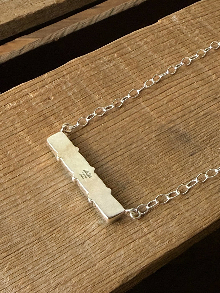 The Navasota Turquoise Bar Necklace by Savannah Sevens western life{&}style rests on a wooden surface, featuring a small engraved design in the center of its sterling silver bar.