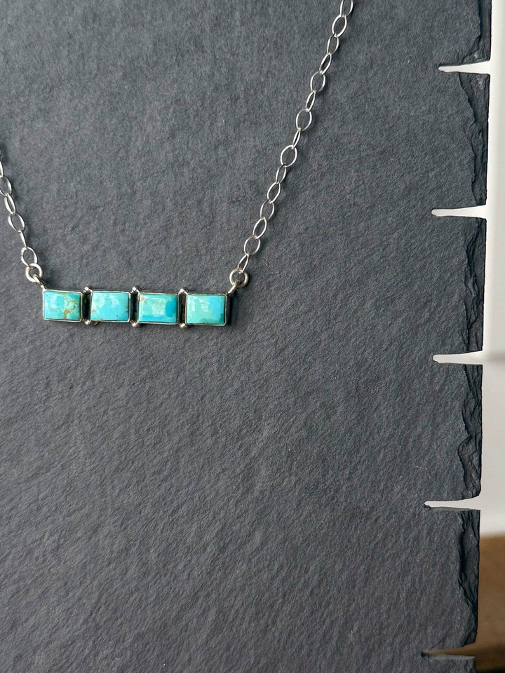The Navasota Turquoise Bar Necklace by Savannah Sevens western life{&}style, featuring four square turquoise stones set in sterling silver, rests on a dark textured background.