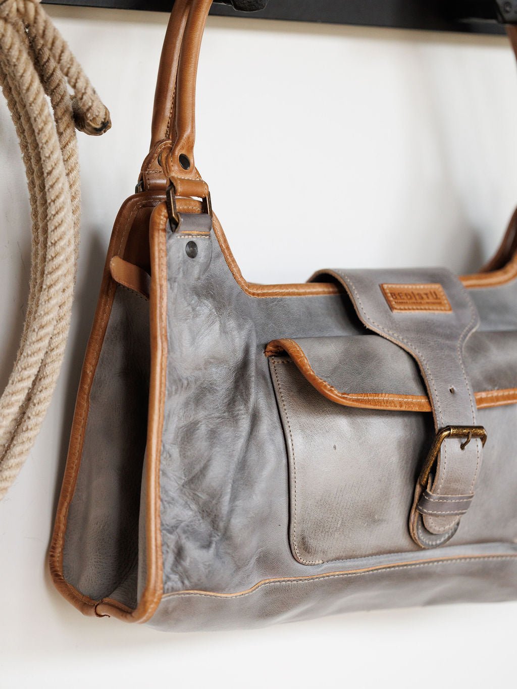 A close-up of the Savannah Sevens western life{&}style Neola Icy Grey Leather Handbag with tan trim and a front pocket, hanging on a hook next to a coiled rope against a white wall. Bag by BedStu. 