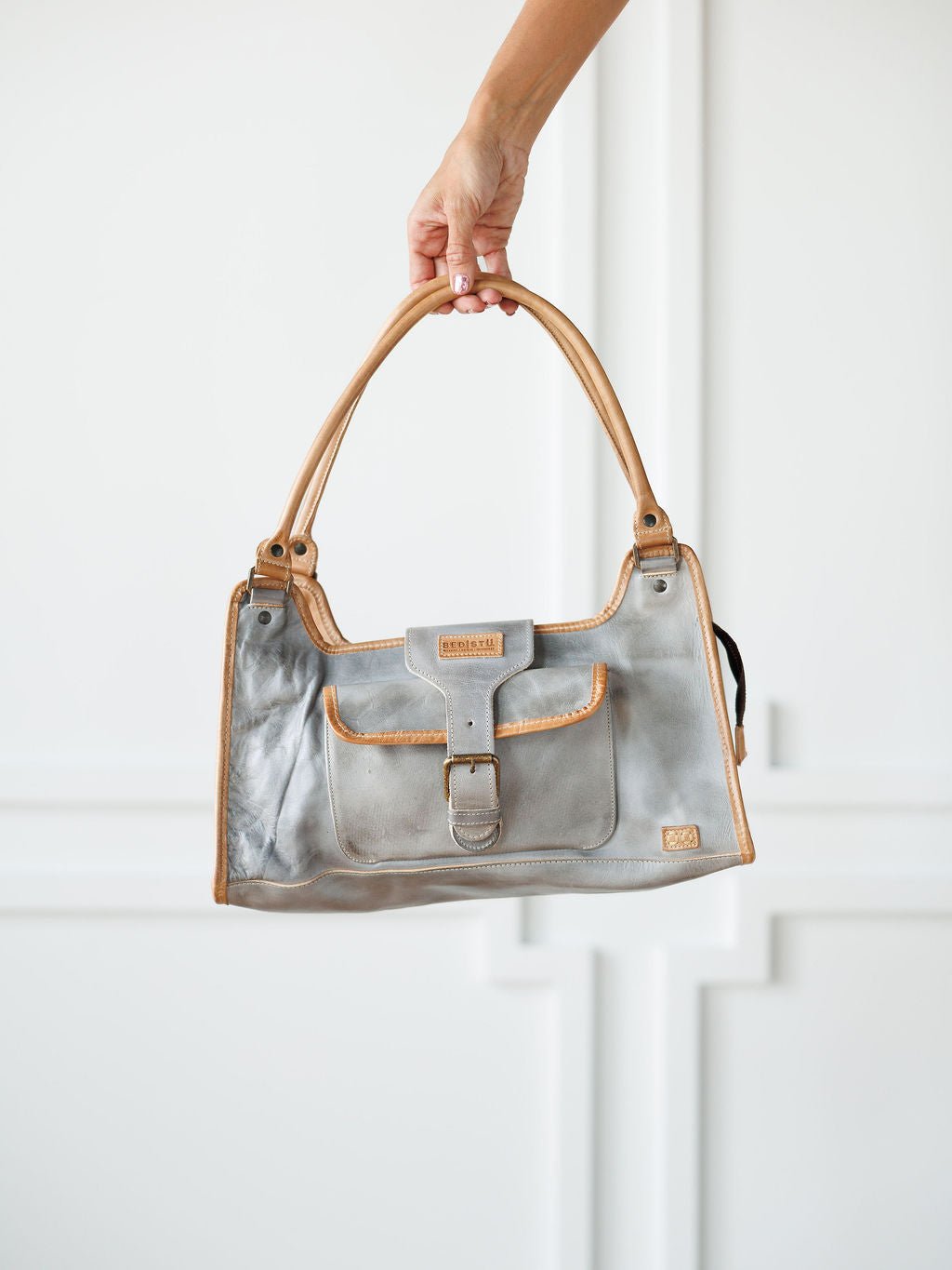 A hand holds the Neola Icy Grey Leather Handbag by Savannah Sevens western life{&}style with tan handles and trim against a white background. This rustic bag features full grain leather, a front buckle pocket, and a small logo patch. Bag by BedStu. 