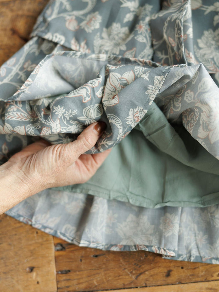 A hand lifts the hem of the Savannah Sevens western life{&}style New Haven Slate Blue Floral Dress, revealing its light green lining. The dress fabric is spread out on a wooden surface.