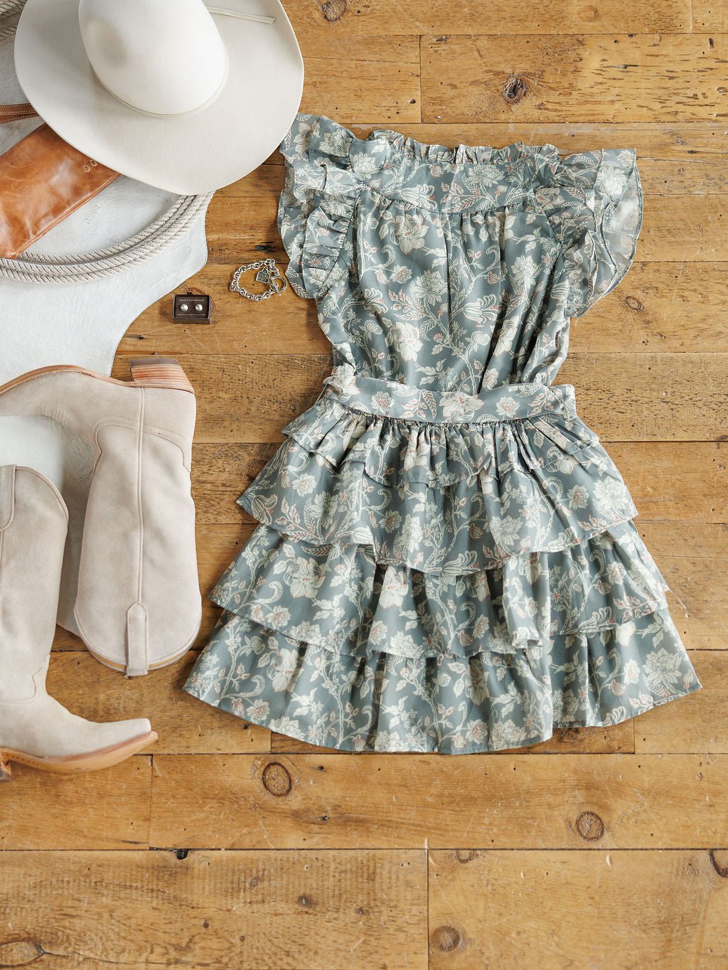 The New Haven Slate Blue Floral Dress by Savannah Sevens western life{&}style is displayed on a wooden floor with cream cowboy boots, a white wide-brim hat, a tan clutch, and a white belt.