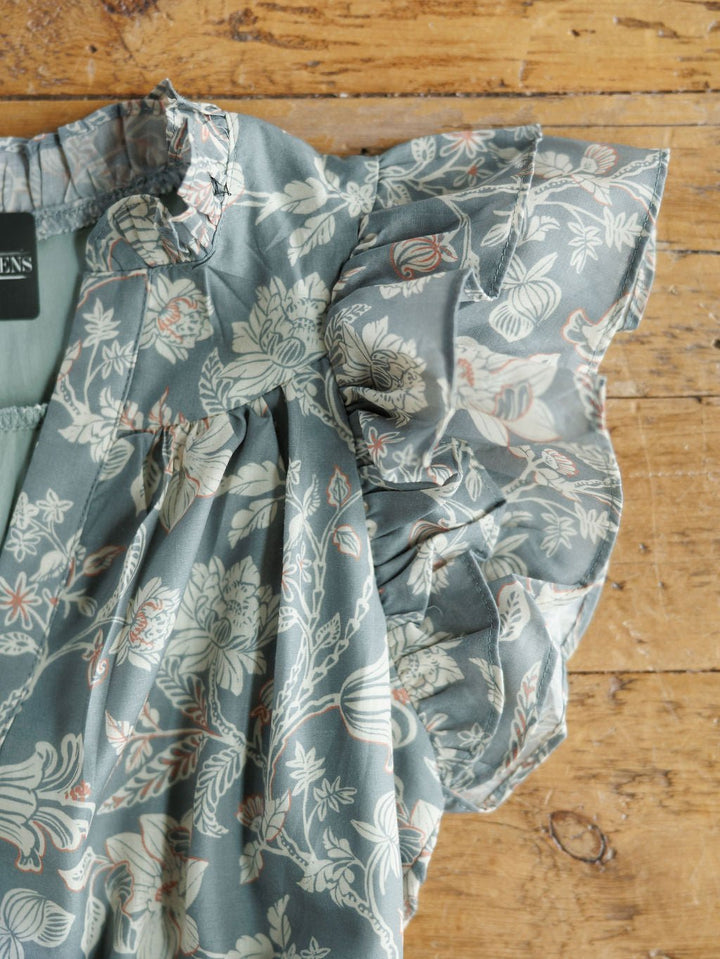 The New Haven Slate Blue Floral Dress by Savannah Sevens western life{&}style features ruffled sleeves and a floral pattern in white, beige, and muted orange, shown on a rustic wooden background.