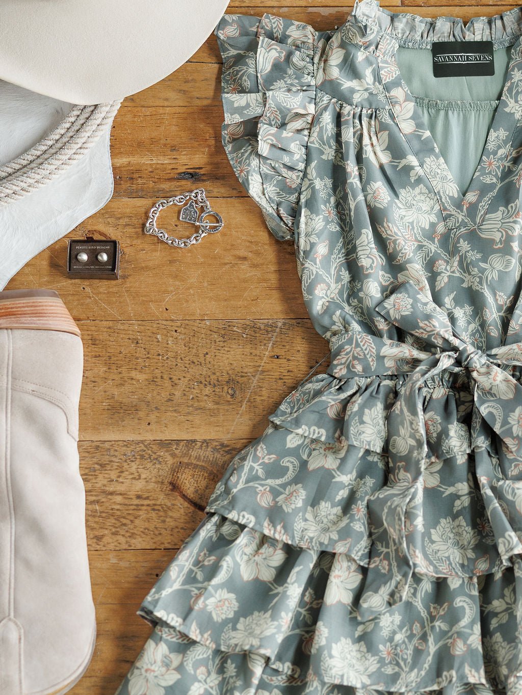 The New Haven Slate Blue Floral Dress by Savannah Sevens western life{&}style is styled on a wooden surface with a beige wide-brim hat, silver bracelet, and light suede knee-high boot.