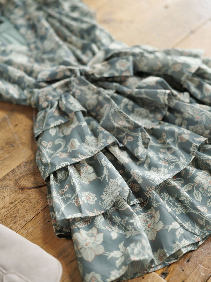 A close-up of the Savannah Sevens western life{&}style New Haven Slate Blue Floral Dress shows its delicate floral and leaf print with layered, tiered ruffles, displayed on a wooden floor.