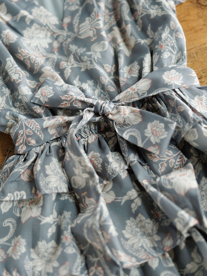 A close-up of the Savannah Sevens western life{&}style New Haven Slate Blue Floral Dress, with white and light pink flowers, a ruffle hem, and tied waist bow, displayed on a wooden surface.