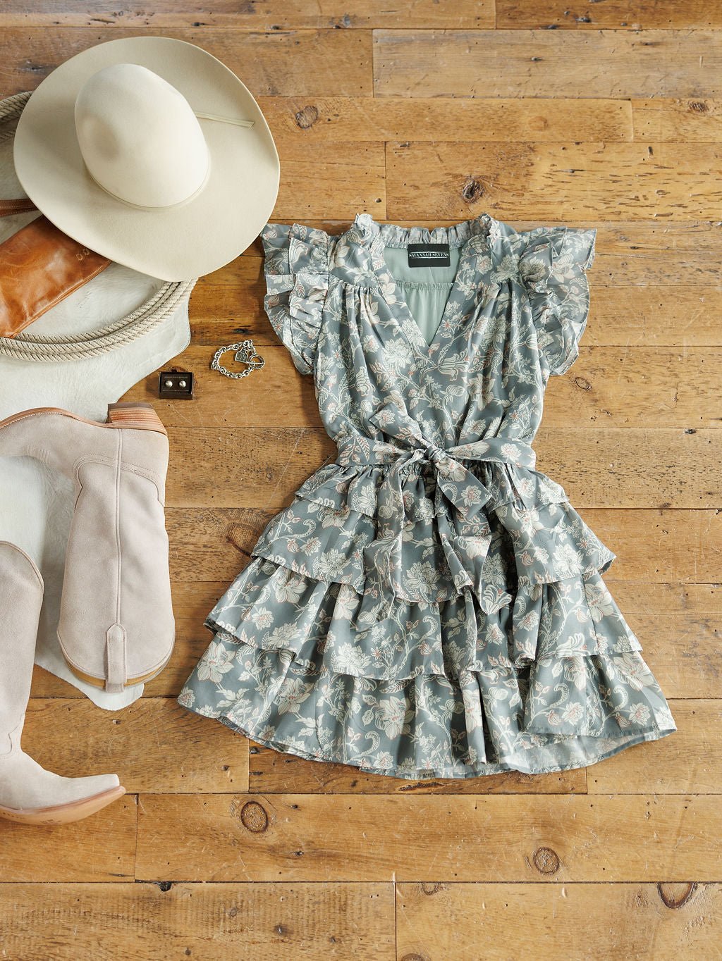 The New Haven Slate Blue Floral Dress by Savannah Sevens western life{&}style is styled in a flat lay with a beige hat, tan clutch, cream boots, bracelet, and earrings for a coordinated look.