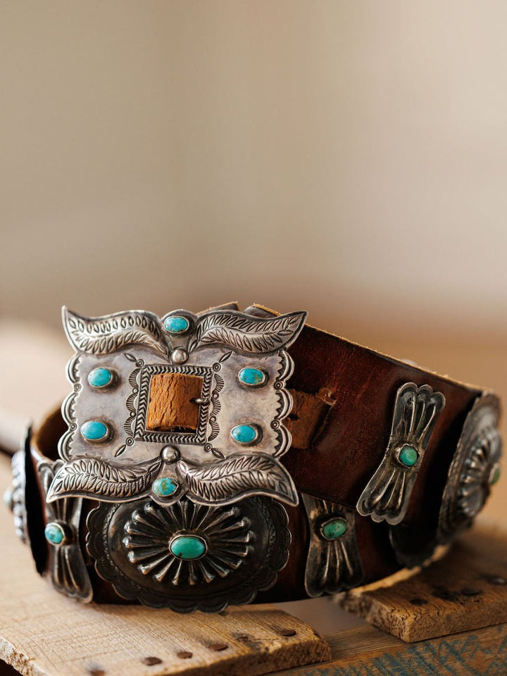 The New Mexico Belt by Savannah Sevens western life{&}style features ornate silver conchos and a large turquoise-accented buckle, beautifully displayed on a rustic wooden surface.