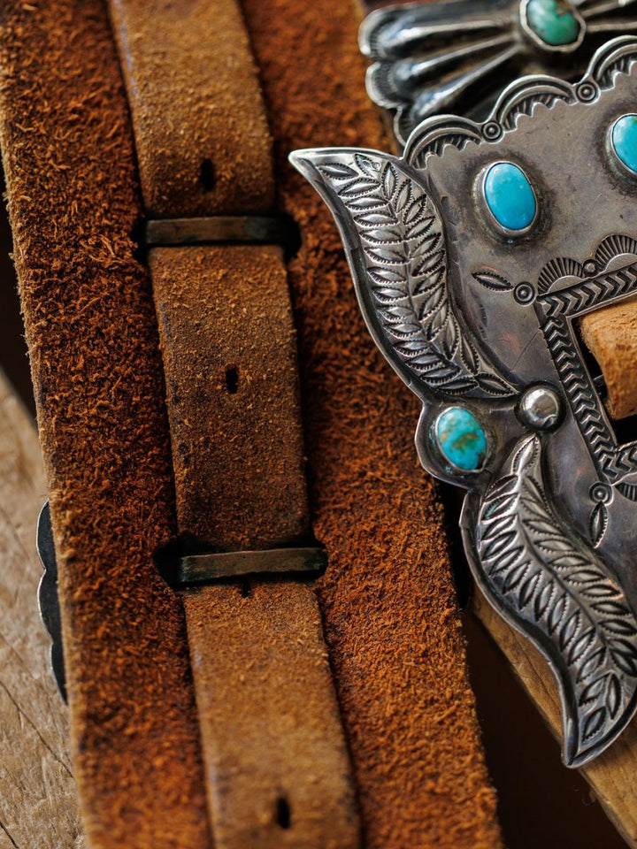 Close-up of the New Mexico Belt by Savannah Sevens western life{&}style, showcasing weathered brown leather and a detailed silver buckle with turquoise stones and engraved leaf motifs. Handmade leather texture and intricate metalwork are visible.