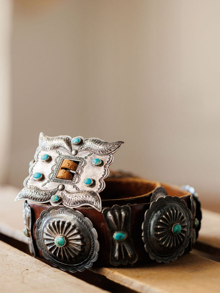 A close-up of the New Mexico Belt by Savannah Sevens western life{&}style, featuring intricate sterling silver and turquoise details, displayed on a wooden surface with a blurred background.