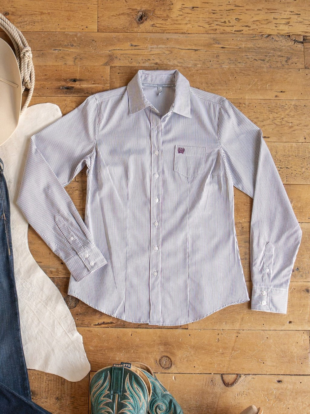 The Newburg Striped Tencel Performance Shirt by Savannah Sevens western life{&}style, featuring light blue and white stripes, is laid flat on a wooden floor alongside a white hat, white fabric, jeans, and turquoise cowboy boots.