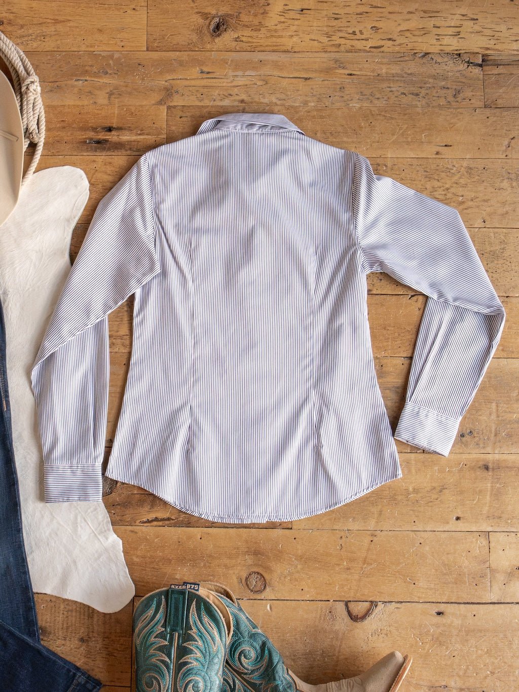 The Newburg Striped Tencel Performance Shirt by Savannah Sevens western life{&}style, a women’s long sleeve blue and white striped button up, is displayed flat on a wooden floor, accompanied by turquoise embroidered cowboy boots.