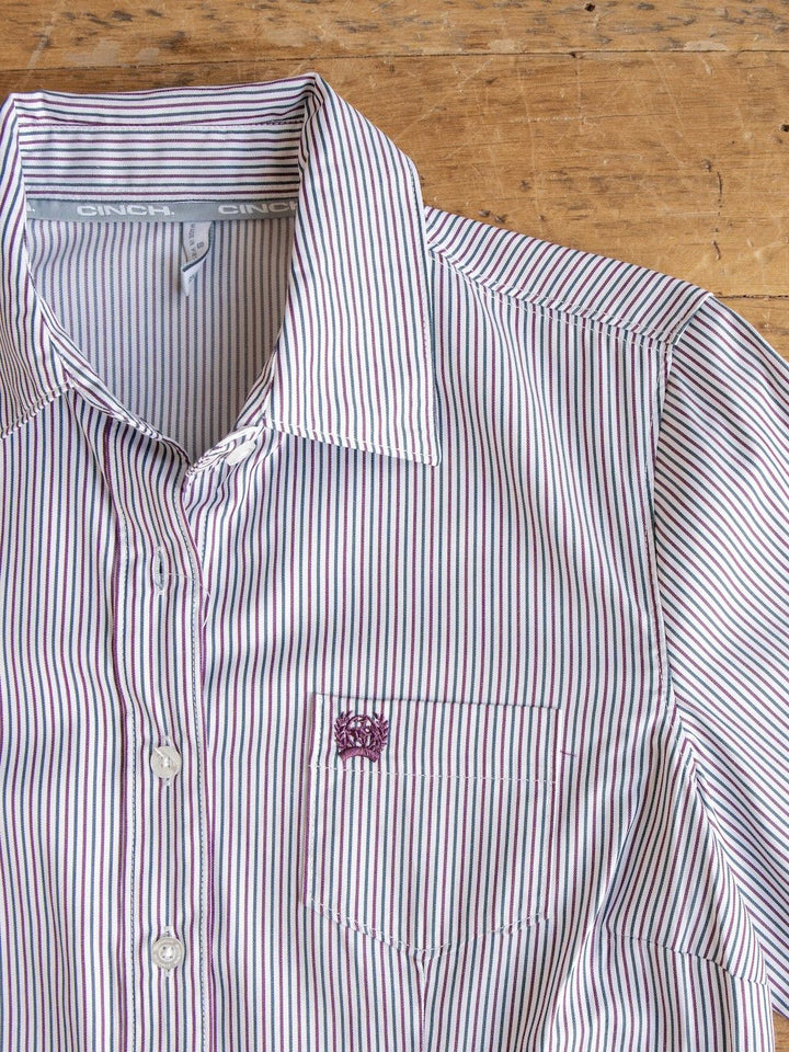 The Newburg Striped Tencel Performance Shirt by Savannah Sevens western life{&}style, a women's long sleeve button up shirt with thin red and blue vertical stripes, a front pocket adorned with a small embroidered emblem, and a folded collar, is laid flat on a wooden surface.