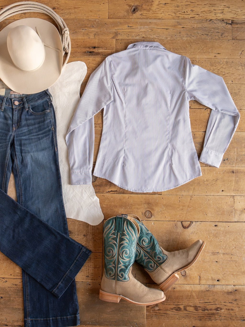 Flat lay of a cowboy hat, blue jeans, tan cowboy boots with blue-green designs, and the Savannah Sevens western life{&}style Newburg Striped Tencel Performance Shirt arranged on a wooden floor.
