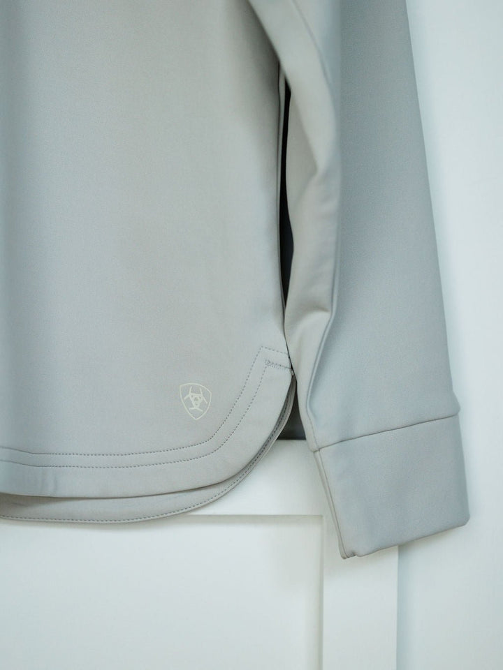 A close-up of the Savannah Sevens western life{&}style Newell Ariat Grey Quarter Zip Pullover hanging on a white door, featuring its moisture-wicking fabric, part of the sleeve, and a small embroidered logo near the hem.