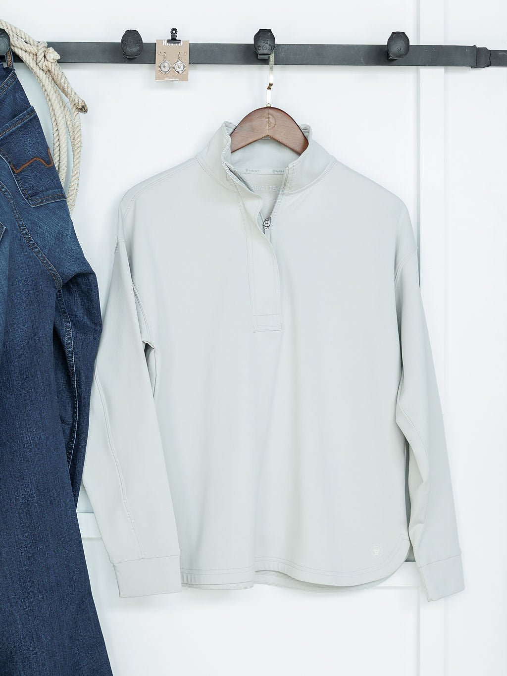 The Newell Ariat Grey Quarter Zip Pullover by Savannah Sevens western life{&}style, made with moisture-wicking fabric, hangs on a wooden hanger from a black wall-mounted rack. Blue jeans and rope are also visible on the rack.
