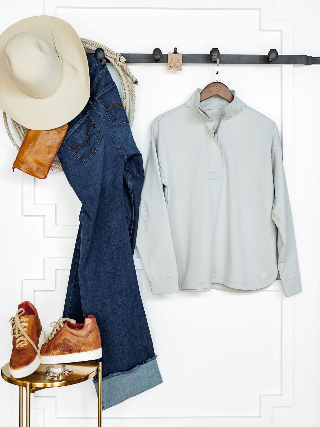 A beige hat, coiled rope, brown leather bag, and blue jeans hang by the Newell Ariat Grey Quarter Zip Pullover from Savannah Sevens western life{&}style. Brown sneakers sit on a gold stool against a white textured wall below.