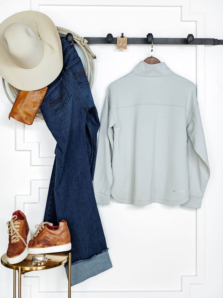 A cream hat, blue jeans, and a tan leather bag hang on a wall rack with the Newell Ariat Grey Quarter Zip Pullover from Savannah Sevens western life{&}style. Tan and white sneakers sit below on a gold stool, while a coiled rope drapes over the rack.
