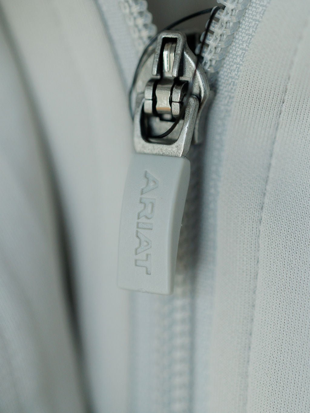 Close-up of the light gray zipper on the Newell Ariat Grey Quarter Zip Pullover by Savannah Sevens western life{&}style, partially open to reveal the fleece lining and textured, moisture-wicking fabric.