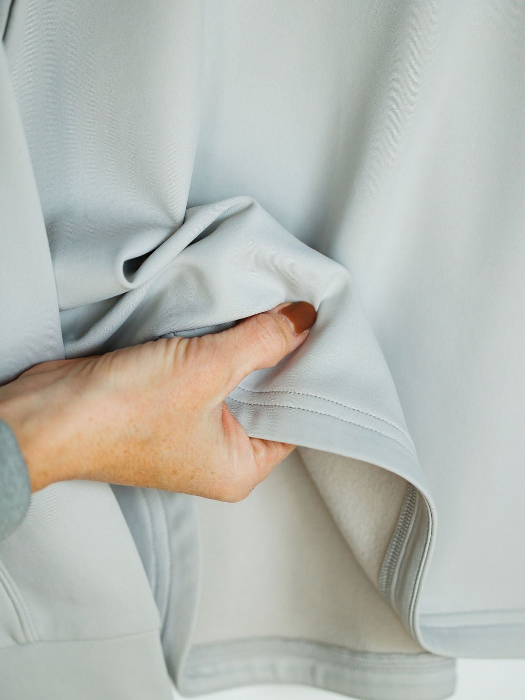 A hand gently pinches and lifts the soft, light gray fabric of the Newell Ariat Grey Quarter Zip Pullover by Savannah Sevens western life{&}style, revealing its smooth texture and fleece-lined, light-colored inner lining.